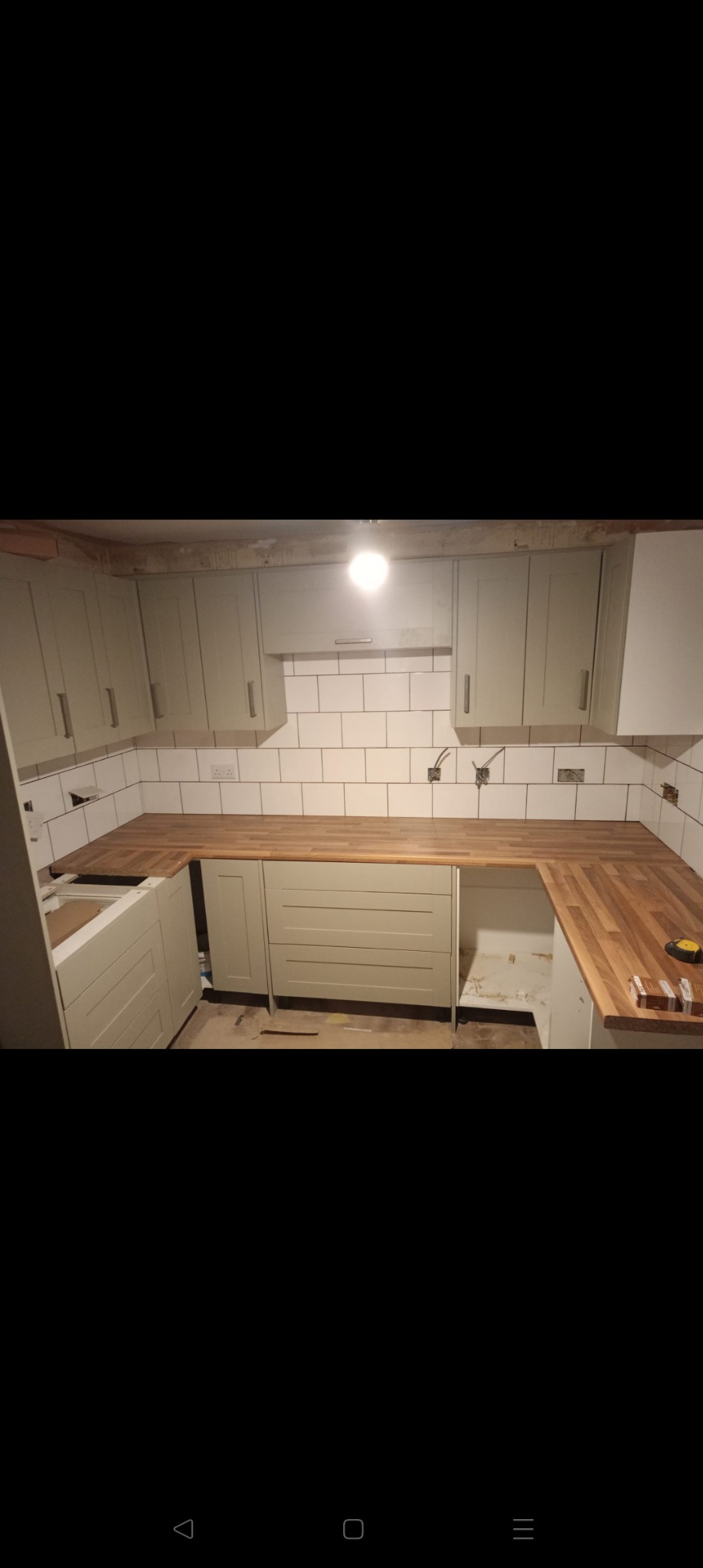 Kuchnia w trakcie montażu: drewniany blat, szare szafki, białe płytki. Widoczne niedokończone instalacje elektryczne i brakujące elementy wykończenia.