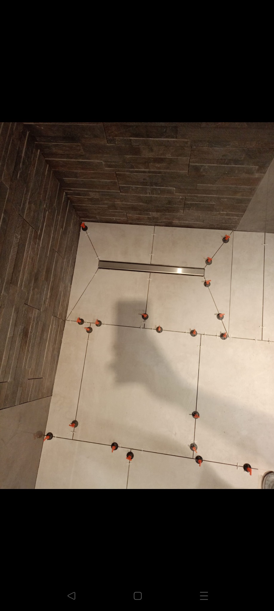 Ułożone płytki podłogowe w łazience z widocznym odpływem liniowym i systemem poziomowania płytek. Ściana wykończona płytkami imitującymi kamień.