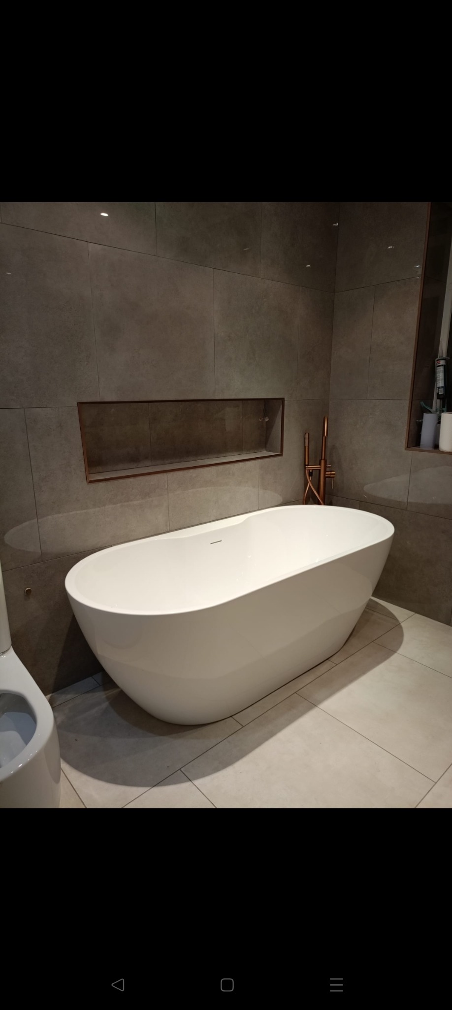 Nowoczesna łazienka z wolnostojącą białą wanną, szarą płytką na ścianach i podłodze oraz miedzianą armaturą. W ścianie wnęka z półką.