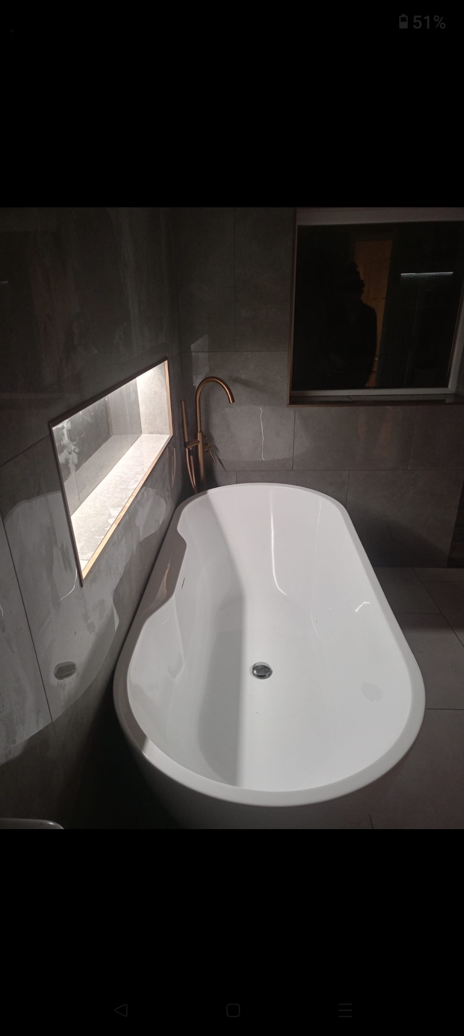 Nowoczesna, biała wanna wolnostojąca z miedzianą armaturą na tle szarych płytek. W ścianie wnęka z podświetleniem. Minimalistyczny design łazienki.