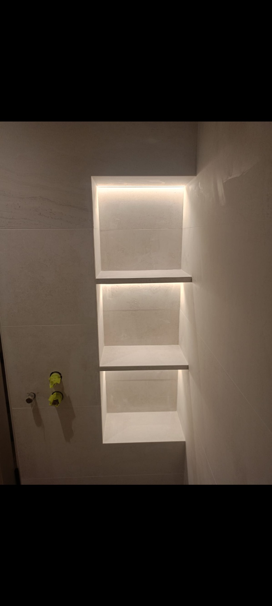 Wykończona wnęka z trzema półkami, podświetlona ledami, w łazience wyłożonej jasnymi płytkami. Widoczne przyłącza wodne.