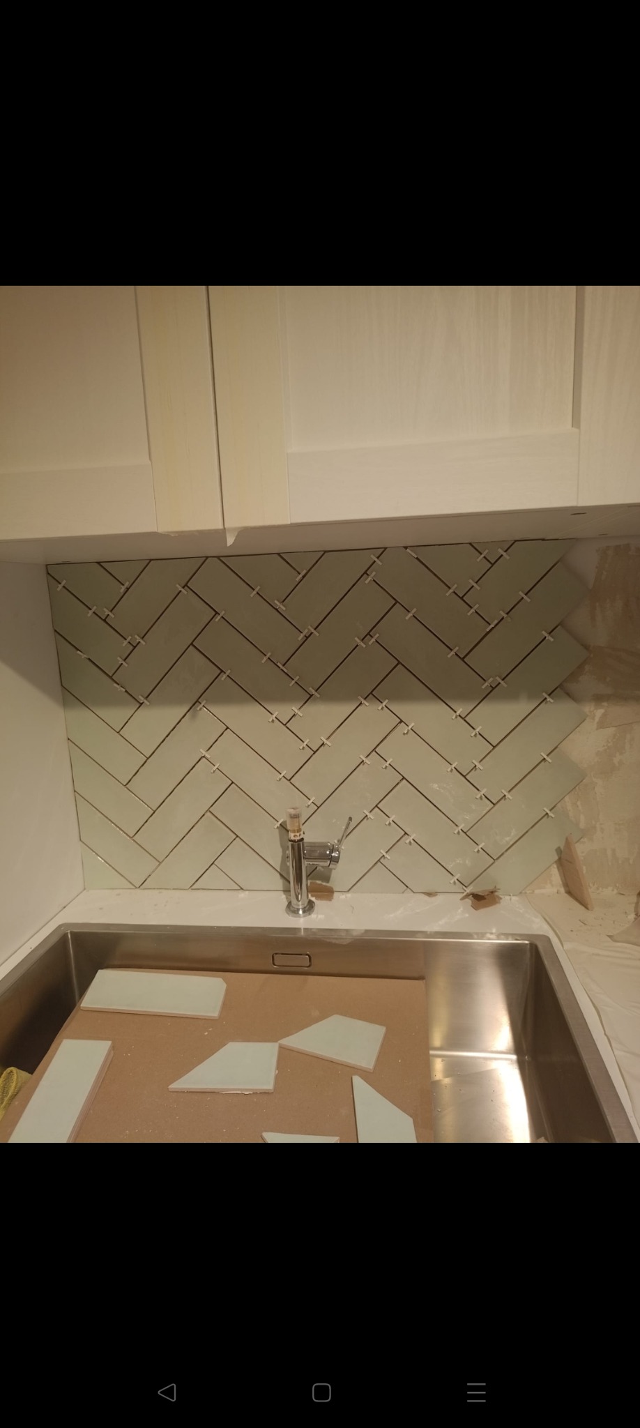 Układanie jasnych płytek w jodełkę nad zlewem w kuchni. Widoczne płytki, fuga i armatura. Częściowo wykończona ściana nad blatem kuchennym.