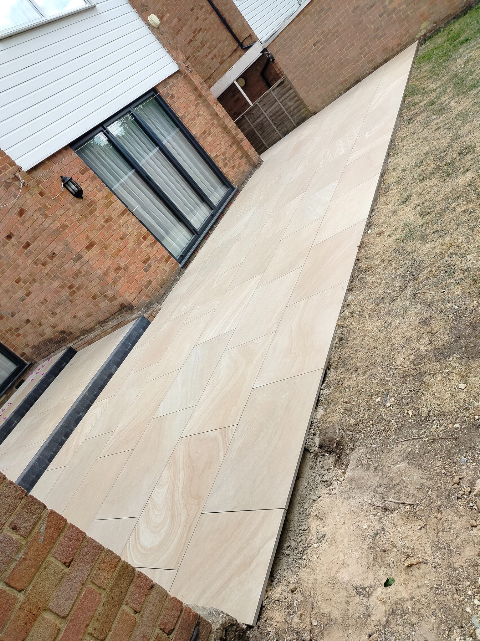 Ułożona, beżowa, prostokątna kostka brukowa tworząca ścieżkę przy ceglanym budynku z oknem. Widoczne obrzeża i trawa. Ujęcie z góry, lekko ukośne.