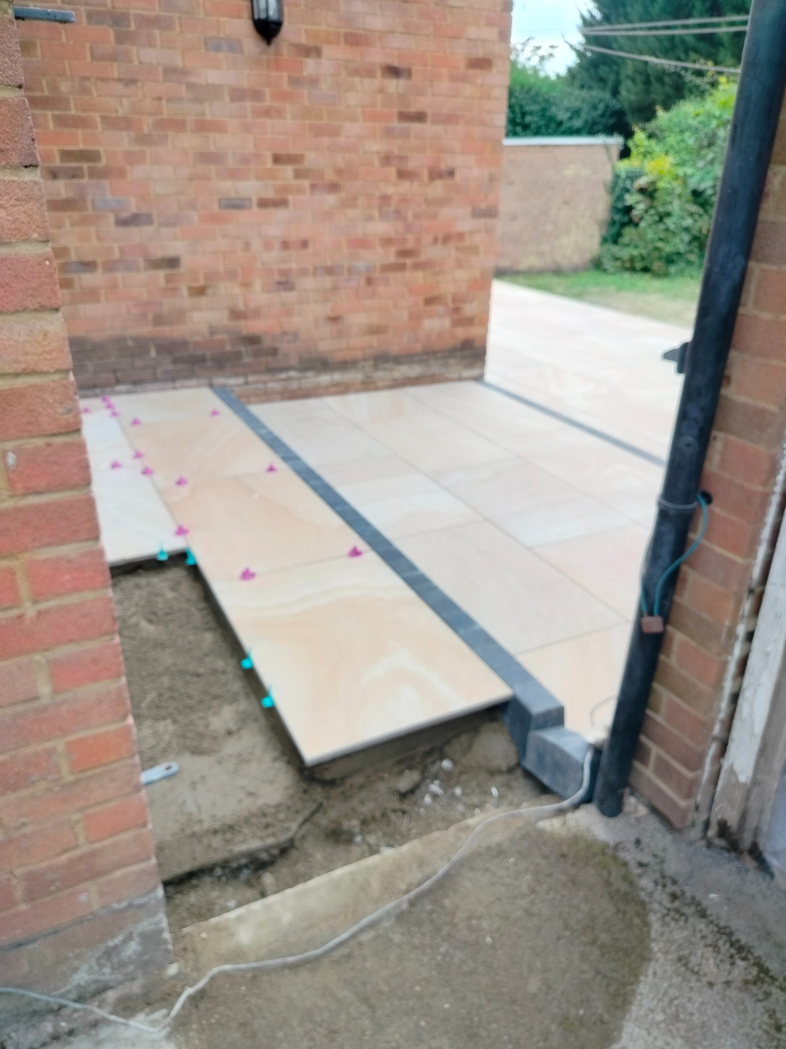 Układanie jasnych płytek na zewnątrz budynku, widoczne elementy mocujące. Fragment muru z cegły w tle. Prace w toku, etap kładzenia płytek.