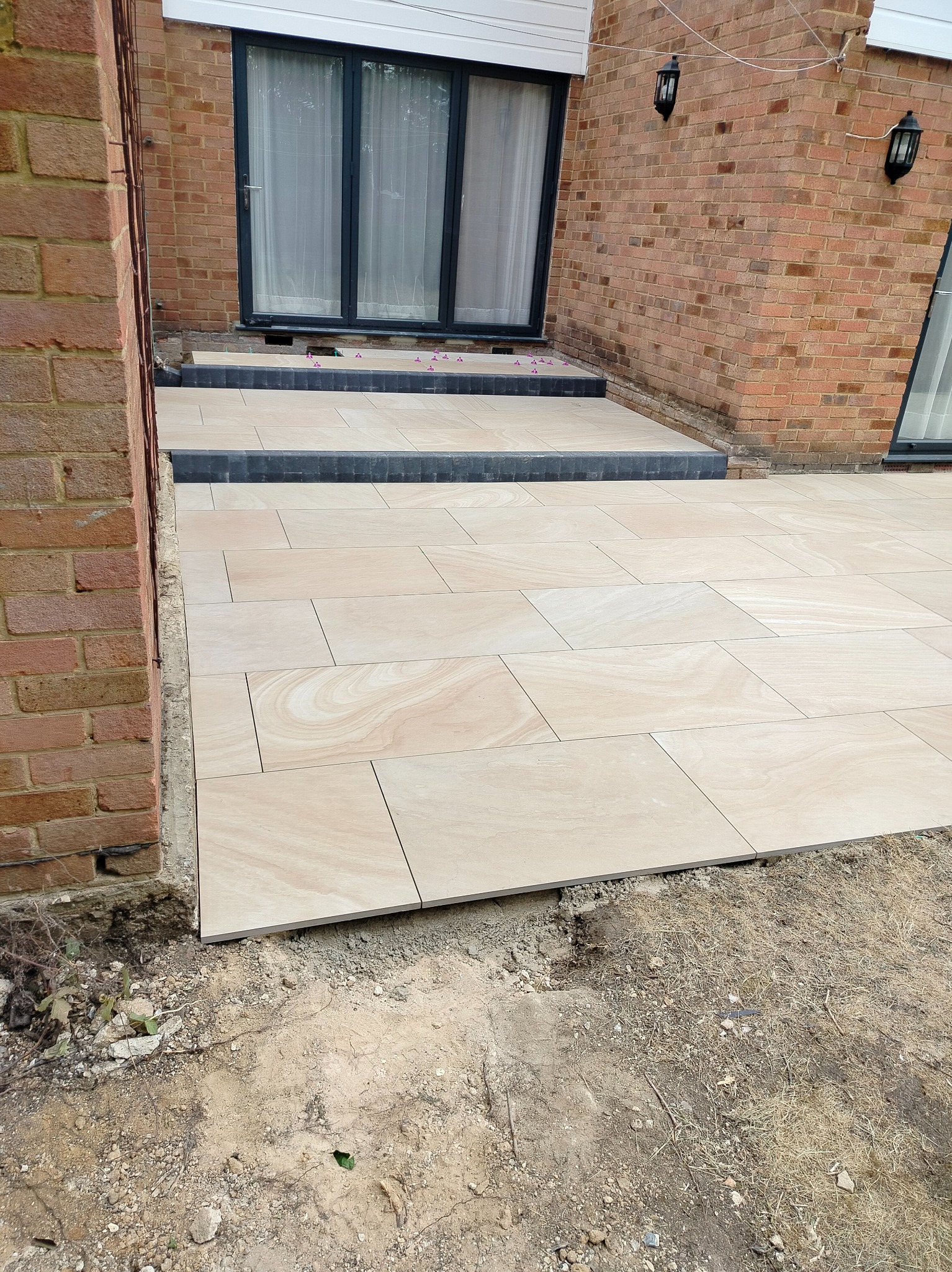 Ułożona, beżowa, prostokątna kostka brukowa na tarasie przy ceglanym domu. Widoczne czarne obrzeża i fragmenty niezakończonych prac ziemnych.