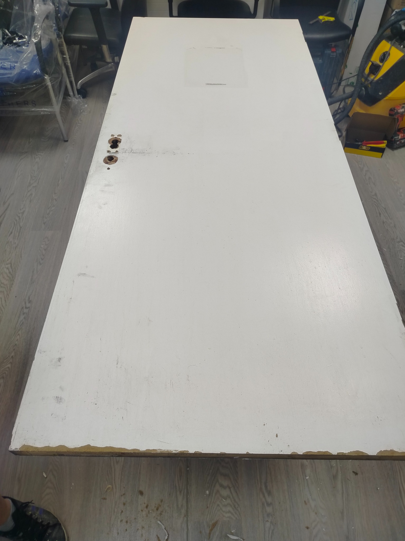 Białe, drewniane drzwi z widocznymi otworami na klamkę i śladami użytkowania, leżące na podłodze. Widoczne uszkodzenia krawędzi i zabrudzenia na powierzchni.