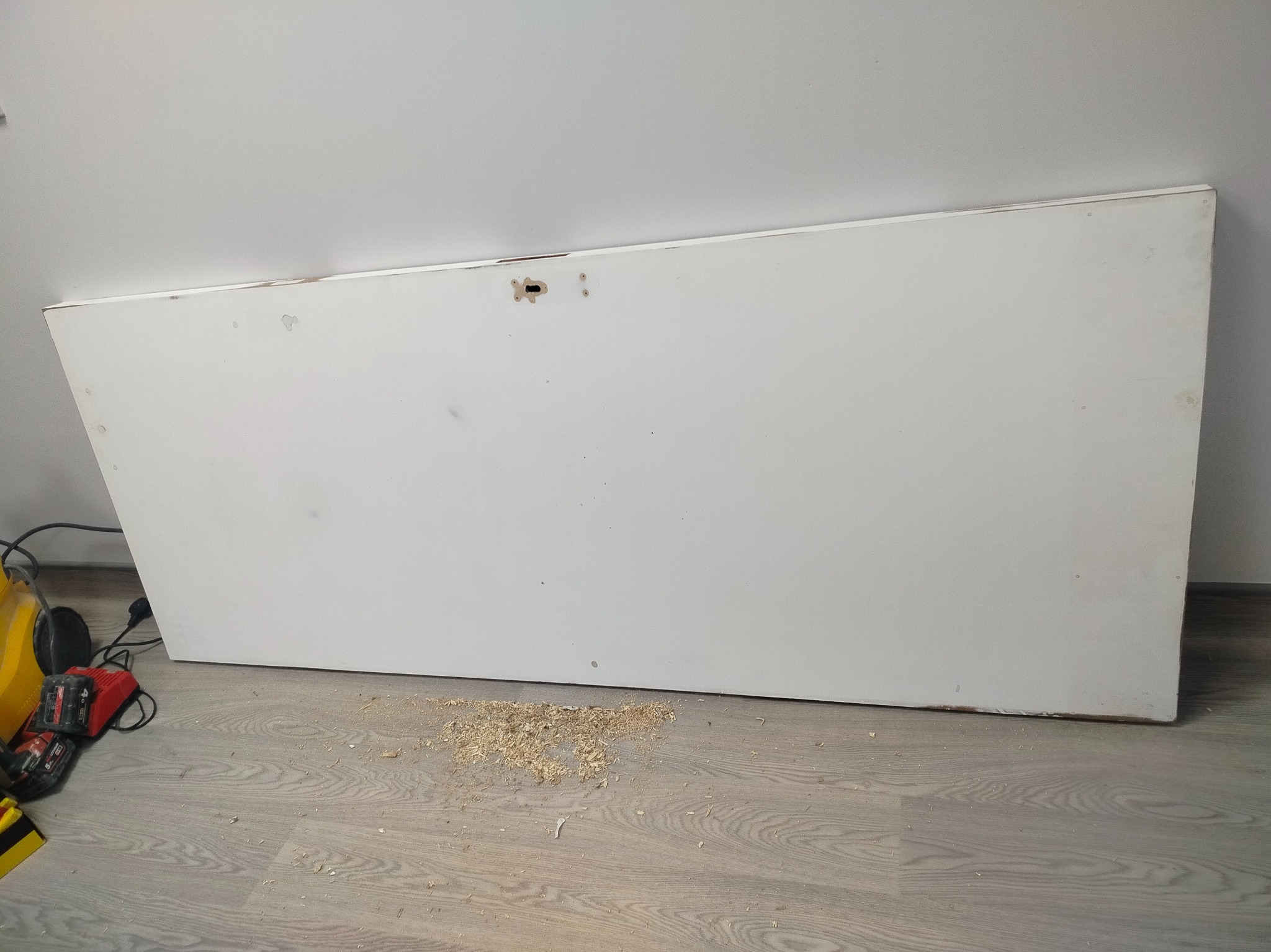 Białe drzwi z widocznymi śladami obróbki stolarskiej leżące na podłodze, obok narzędzia i wióry. Praca w toku, przygotowanie do montażu lub renowacji.