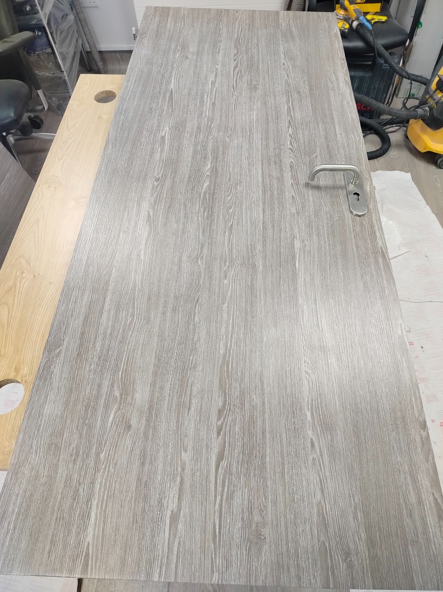 Drzwi wewnętrzne z szarą okleiną drewnopodobną, leżące na blacie roboczym. Widoczny srebrny uchwyt i zamek. W tle narzędzia i odkurzacz.