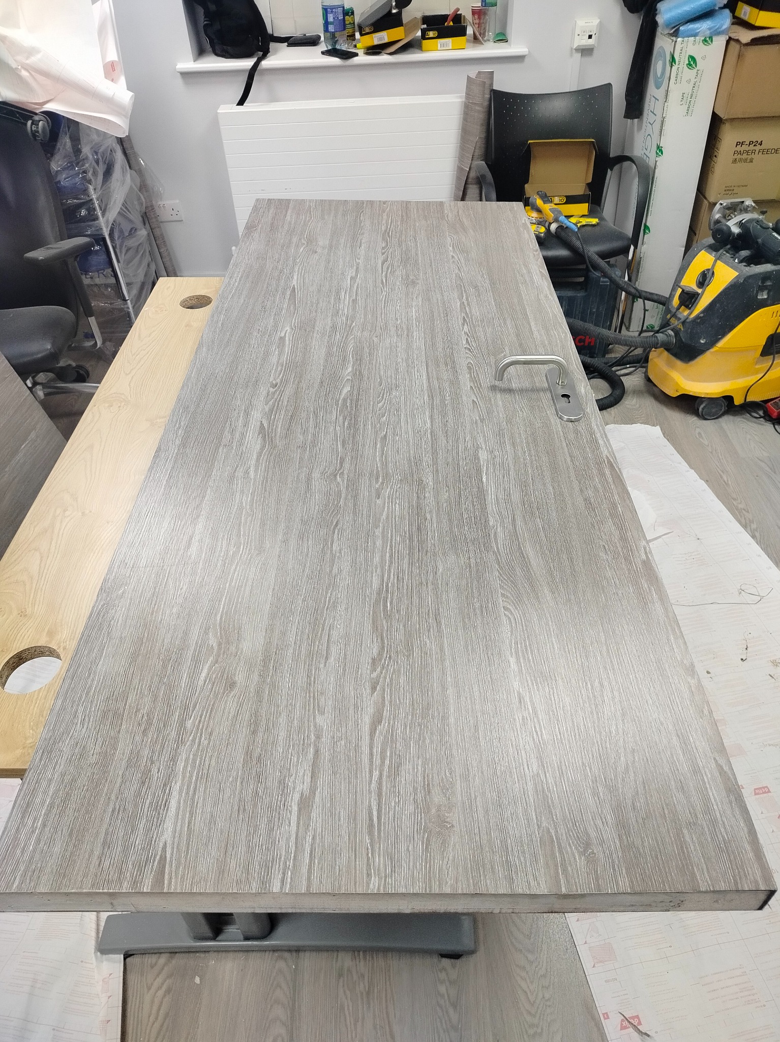 Blat biurka z szarą okleiną drewnopodobną, z zamontowanym uchwytem, leżący na stole roboczym. W tle widoczne narzędzia i akcesoria.