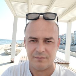 CHANCE FOR PURITY Volodymyr Dzhogola - Mężczyzna w białej koszulce i okularach na głowie, na tle nadmorskiej promenady z białymi ławkami i pergolą. Widok na morze i budynki w oddali.