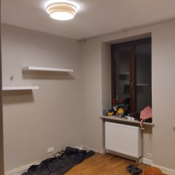 Pokój po malowaniu: świeże, beżowe ściany, białe półki i grzejnik. Podłoga zabezpieczona folią malarską, widoczne narzędzia. Nowoczesne oświetlenie sufitowe.