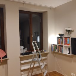 Pokój w trakcie odświeżania: drabina stoi przy oknie z narzędziami, widoczne ślady prac malarskich na ścianie i suficie. Na ścianie półka z książkami i dekoracjami.