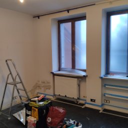 Pokój w trakcie przygotowania do malowania: zabezpieczone okna folią, ściany z widocznymi śladami po naprawach, drabina i akcesoria malarskie na podłodze.