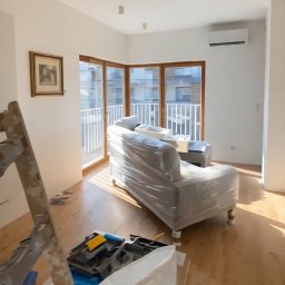 Jasne wnętrze w trakcie malowania: zabezpieczony folią narożnik, drabina malarska, puszka farby. Widoczne ślady farby na podłodze i ścianach. Słoneczne światło wpada przez okna.