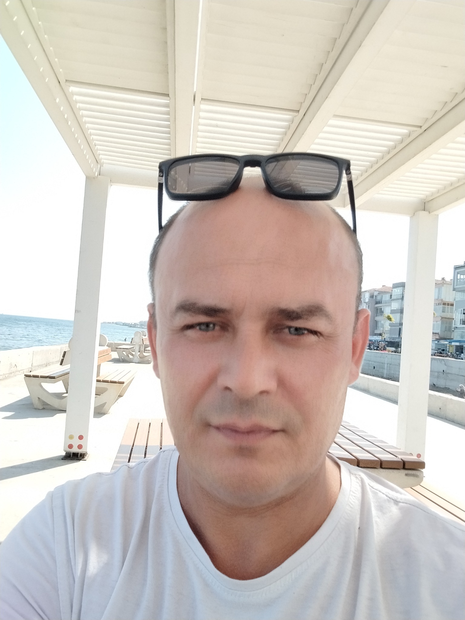 Mężczyzna w białej koszulce i okularach na głowie, na tle nadmorskiej promenady z białymi ławkami i pergolą. Widok na morze i budynki w oddali.