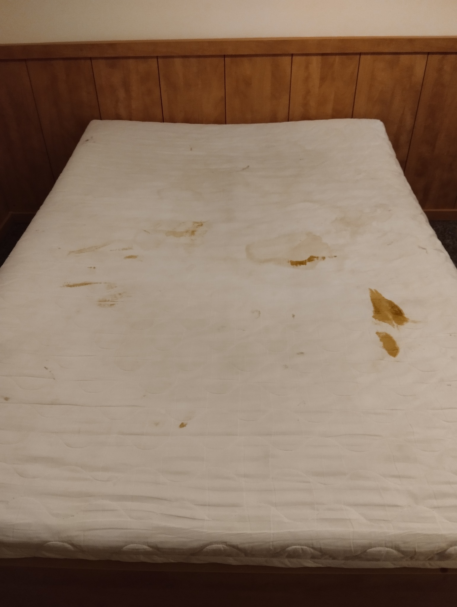 Zabrudzony materac z widocznymi plamami na białym poszyciu, umieszczony na drewnianym łóżku z wezgłowiem. Widoczne ślady użytkowania i konieczność odświeżenia.