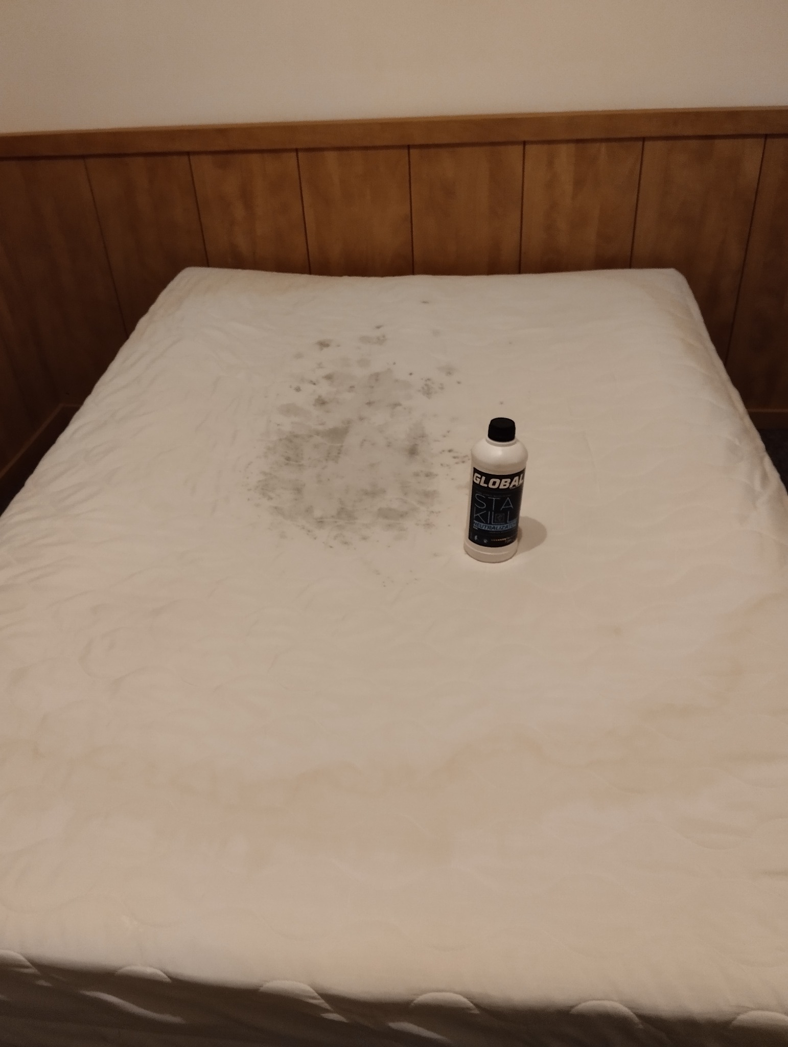 Zabrudzony materac z plamami przed czyszczeniem, obok butelka środka czyszczącego. Widok z góry na materac z wyraźnymi plamami i produktem do czyszczenia.