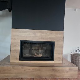 Gabriel Mokwa - Nowoczesny kominek z obudową z paneli drewnopodobnych i czarną ścianą, uzupełniony o kosz na drewno. Element wykończenia wnętrza z widocznymi schodami.
