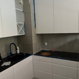 Gabriel Mokwa - Kuchnia w trakcie wykańczania: białe szafki, czarny blat i szare, pionowe płytki w kształcie rombów na ścianie. Widoczne krzyżyki dystansowe między płytkami.