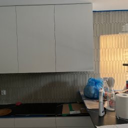 Gabriel Mokwa - Montaż płytek w kuchni z białymi szafkami. Widoczne narzędzia i materiały budowlane. Płytki ułożone w pionowy wzór, zabezpieczone taśmą malarską.