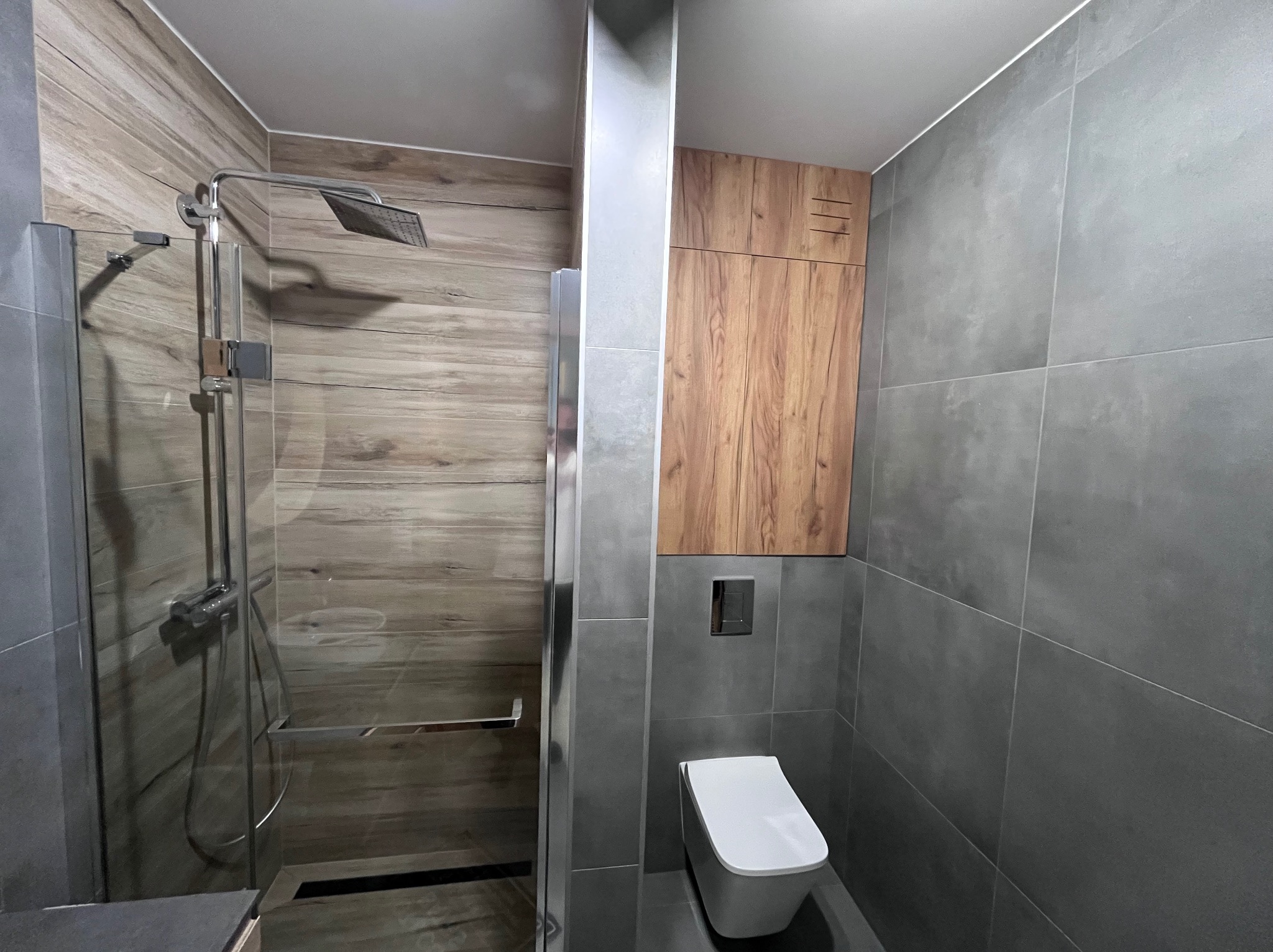 Nowoczesna łazienka z kabiną prysznicową typu walk-in, szarą glazurą i drewnianą szafką nad toaletą. Minimalistyczny design, oświetlenie punktowe.