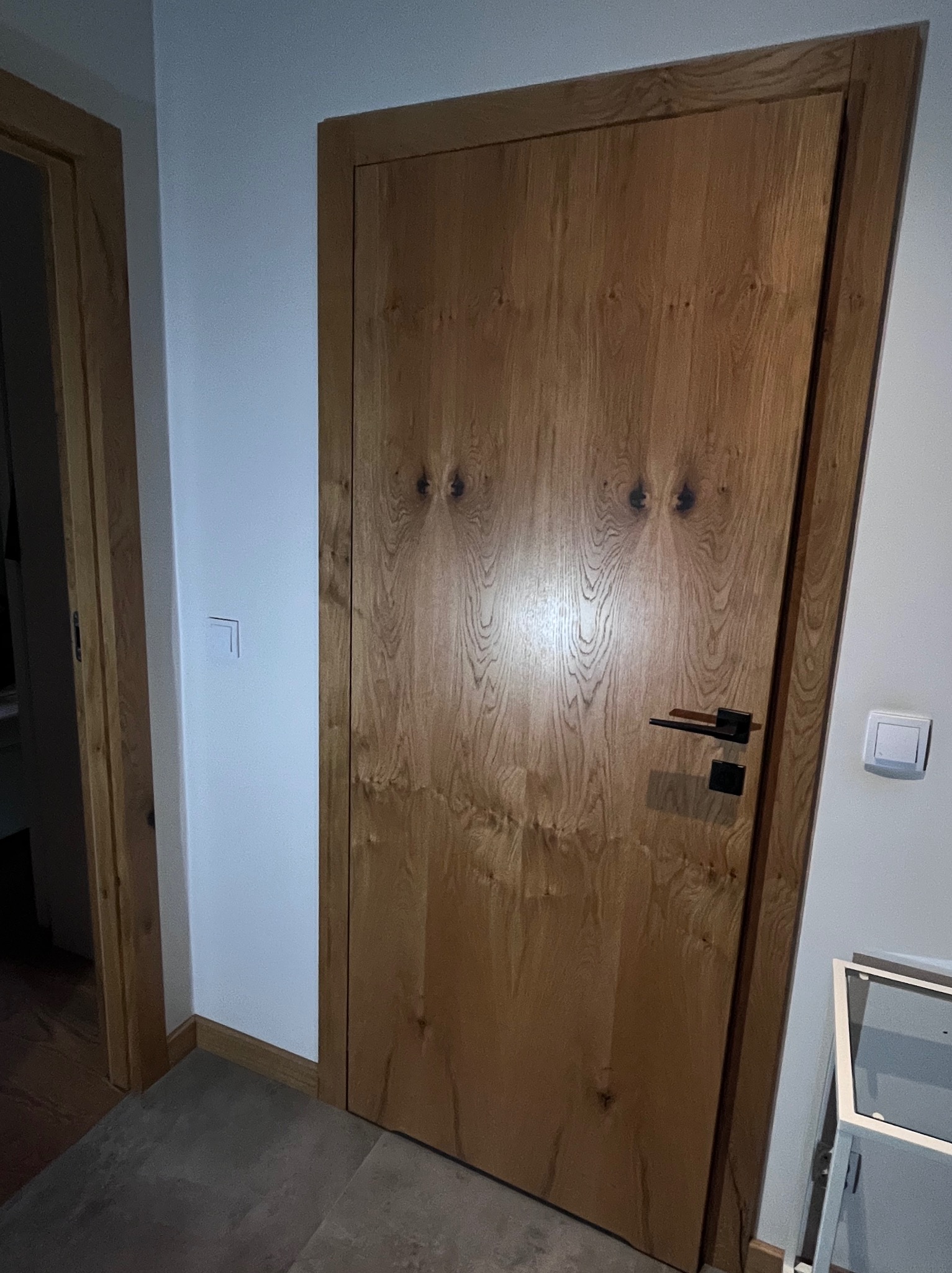Dębowa, fornirowana, wewnętrzna stolarka drzwiowa z czarną klamką, wkomponowana w minimalistyczne wnętrze. Widoczny fragment podłogi i ściany z włącznikiem.