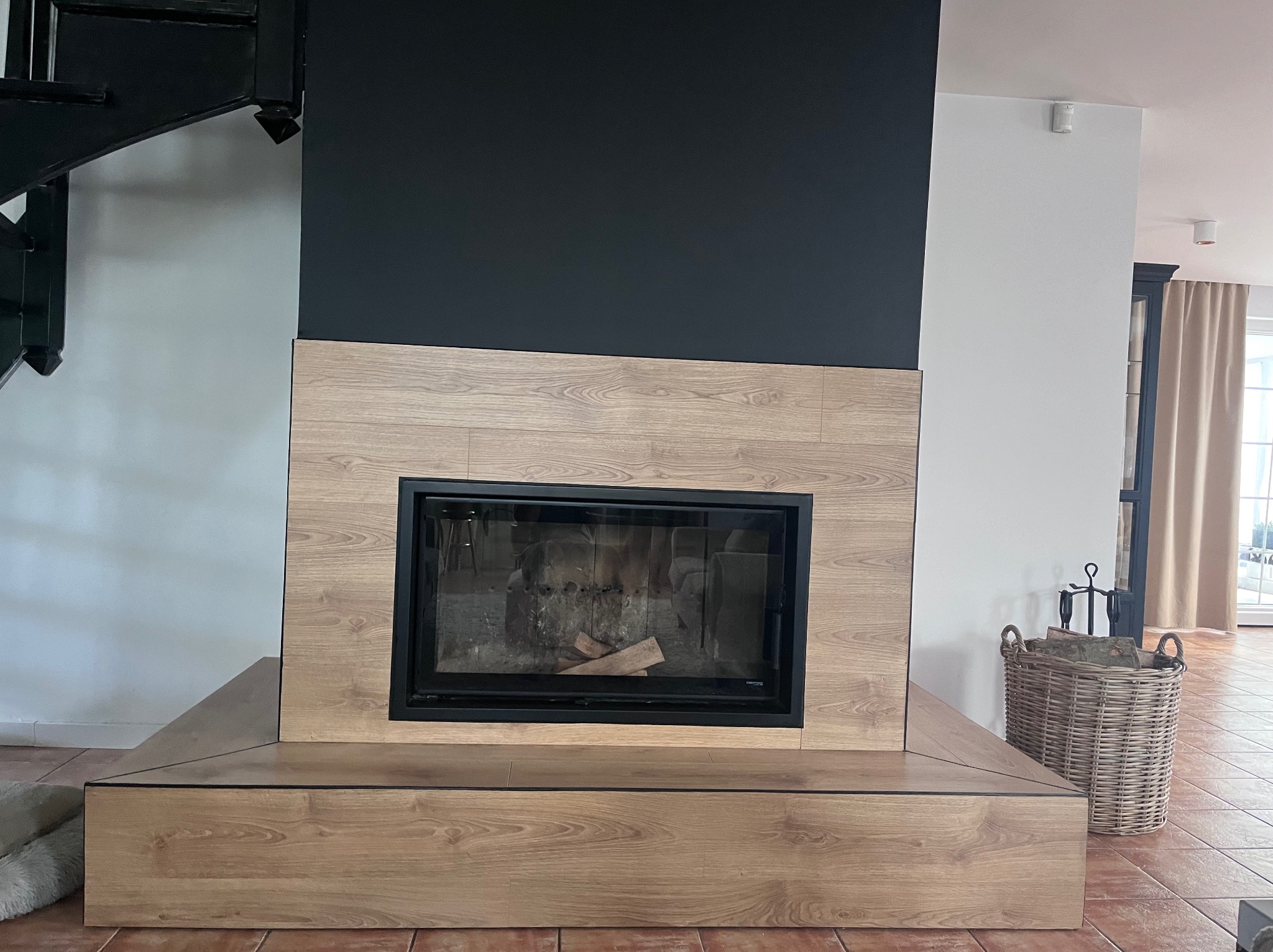 Nowoczesny kominek z obudową z paneli drewnopodobnych i czarną ścianą, uzupełniony o kosz na drewno. Element wykończenia wnętrza z widocznymi schodami.