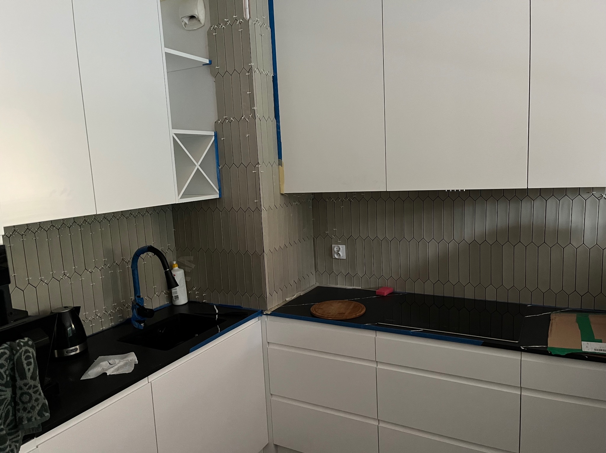 Kuchnia w trakcie wykańczania: białe szafki, czarny blat i szare, pionowe płytki w kształcie rombów na ścianie. Widoczne krzyżyki dystansowe między płytkami.