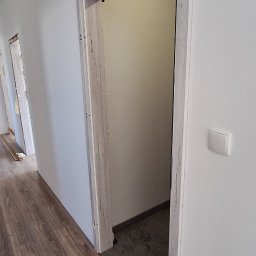 LT. Tom-Bud - Nowo zamontowane, jasne ościeżnice drzwiowe z efektem drewna, widoczne na tle białych ścian i paneli podłogowych. W tle widoczne kolejne drzwi.