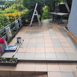 LT. Tom-Bud - Malowanie elewacji domu: drabina oparta o ścianę, narzędzia malarskie na tarasie z płytkami, widok na ogród i jezioro w tle. Świeżo pomalowana ściana na szaro.