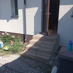 LT. Tom-Bud - Zewnętrzne schody wejściowe wyłożone płytkami imitującymi drewno, prowadzące do otwartych, brązowych drzwi wejściowych. Obok roślinność i elementy otoczenia domu.