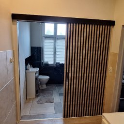 LT. Tom-Bud - Nowoczesne drzwi przesuwne lamelowe w łazience z czarnym marmurem, widok z kuchni. Jasne płytki na ścianach, minimalistyczny design.