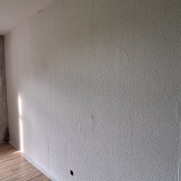 LT. Tom-Bud - Pokój po częściowym malowaniu ścian: jedna ściana biała z fakturą, druga szara z efektem dekoracyjnym. Widoczne podłączenia do lamp i podłoga z paneli.