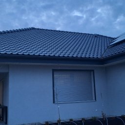 LT. Tom-Bud - Szary dom jednorodzinny z ciemną dachówką i roletami w oknach, sfotografowany o zmierzchu. Widoczny fragment tarasu i rośliny w donicach.