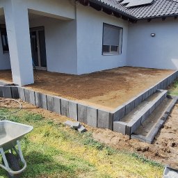 LT. Tom-Bud - Budowa tarasu z betonowych elementów przy domu jednorodzinnym. Widoczna taczka i narzędzia. Etap przygotowania podłoża i układania obrzeży. Słoneczny dzień.