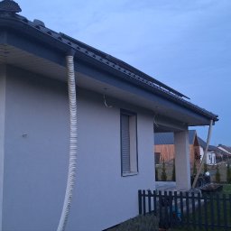 LT. Tom-Bud - Elewacja domu jednorodzinnego po malowaniu, z widoczną rurą spustową i oknem z żaluzjami. Świeża, jasna farba, ciemna dachówka i elementy wykończeniowe.