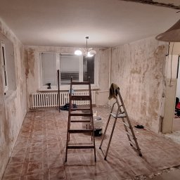 LT. Tom-Bud - Pokój w trakcie remontu: ściany przygotowane do malowania, widoczne drabiny, narzędzia i resztki starej farby. Podłoga częściowo odsłonięta, widoczne ślady po kleju.