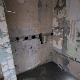 LT. Tom-Bud - Wnętrze w trakcie remontu: oświetlona podłoga z wylewką, ściany z widocznymi śladami usuniętych płytek i instalacji. Widoczne otwory w ścianie i fragmenty starej farby.