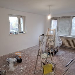 LT. Tom-Bud - Pokój po malowaniu ścian na biało, widoczne narzędzia malarskie, drabina i zabezpieczone okno folią. Podłoga częściowo zakryta farbą. Gniazdko elektryczne bez osłony.