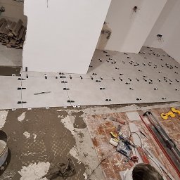 LT. Tom-Bud - Układanie jasnych płytek podłogowych z użyciem klipsów poziomujących. Widoczne narzędzia glazurnicze i wiadro z zaprawą. Fragment grzejnika przy oknie.