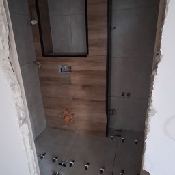 LT. Tom-Bud - Łazienka w trakcie remontu: szare płytki na ścianach i podłodze, drewnopodobne panele na ścianie, widoczne elementy instalacji i krzyżyki glazurnicze.