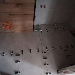 LT. Tom-Bud - Układanie płytek w łazience z użyciem klipsów poziomujących. Częściowo wykończona ściana i sufit z widoczną instalacją hydrauliczną.