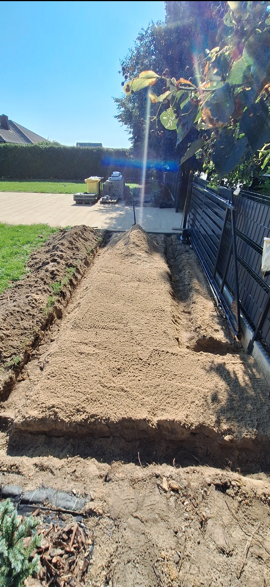 Przygotowanie podłoża pod kostkę brukową: wyrównany piasek w wykopie, widoczne obrzeża i fragment ogrodzenia. Słoneczny dzień, błękitne niebo.