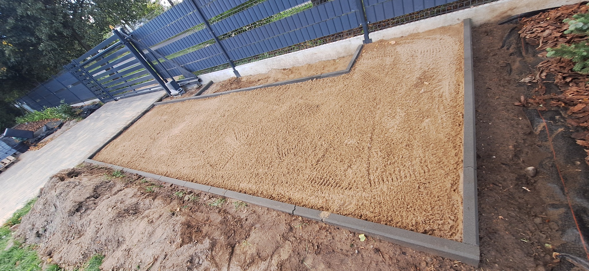 Przygotowany teren pod kostkę brukową z obrzeżami, widoczny fragment chodnika i ogrodzenie panelowe w tle. Piasek wyrównany, gotowy do dalszych prac.