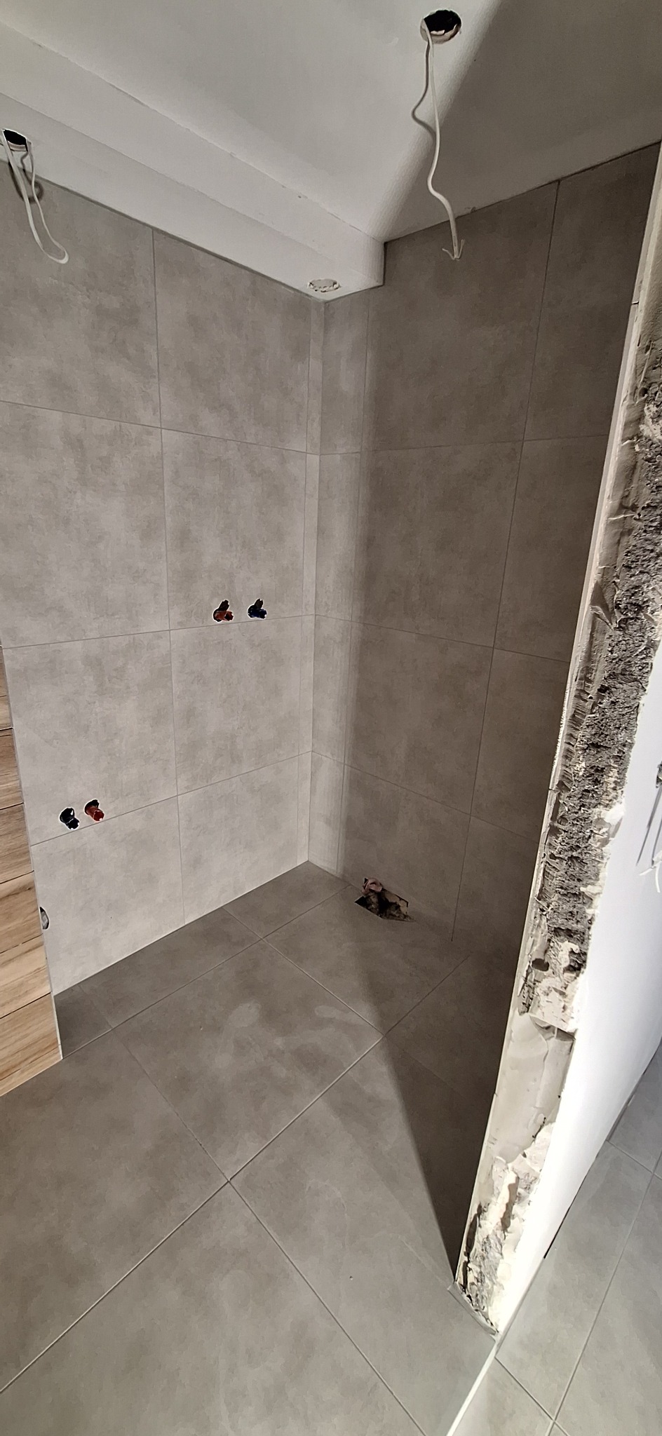 Wykończona łazienka z szarymi płytkami, widoczne przyłącza wodne i instalacja elektryczna. Fragment ściany z widocznym uszkodzeniem, drzwi z drewnianym wykończeniem.