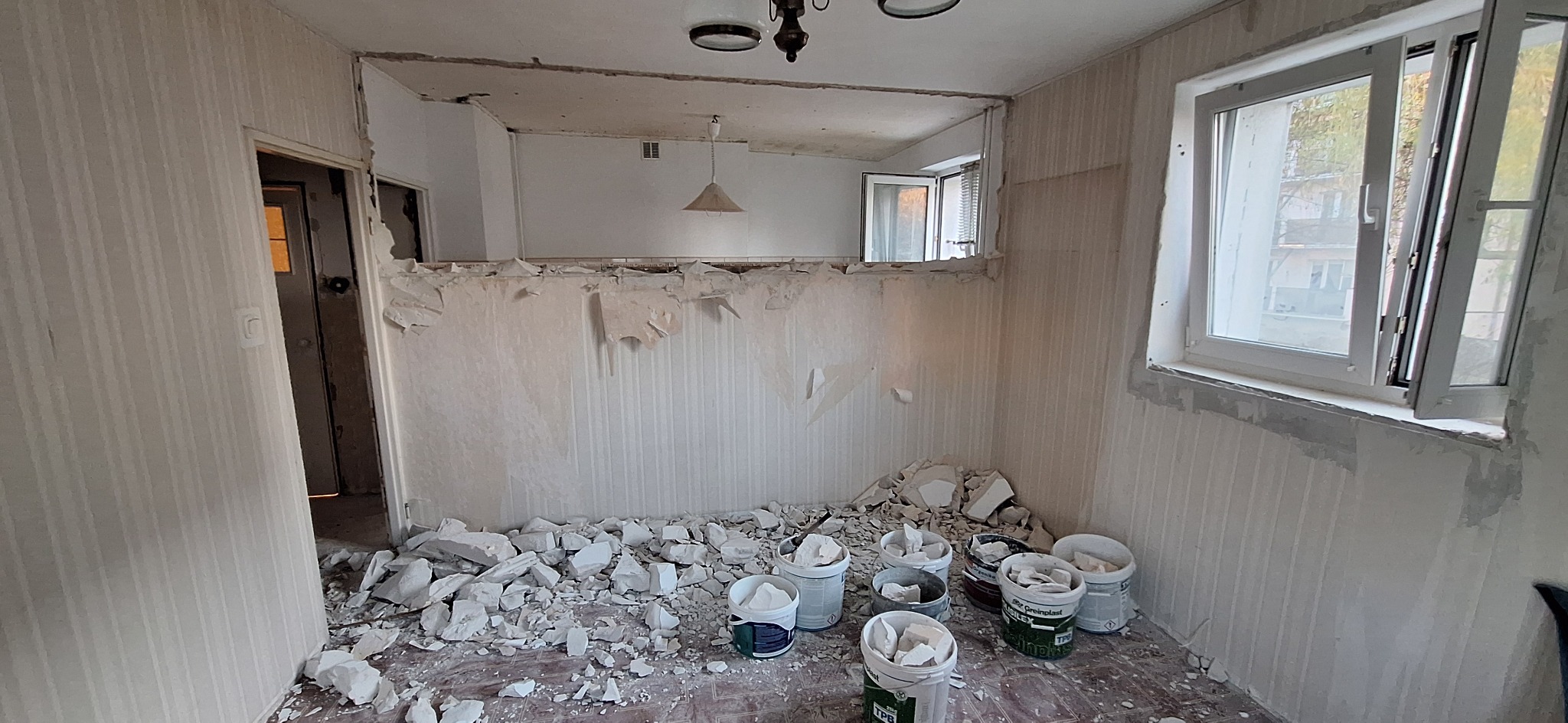 Pokój w trakcie demontażu ściany działowej, widoczne resztki tapety, gruz i wiadra z zaprawą. Okno z otwartym skrzydłem w tle.