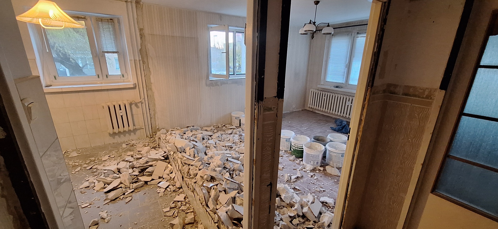 Pokój w trakcie demontażu płytek i przygotowania do remontu. Widoczne gruz, wiadra z farbą i odsłonięte ściany. Ujęcie z perspektywy przejścia między pomieszczeniami.