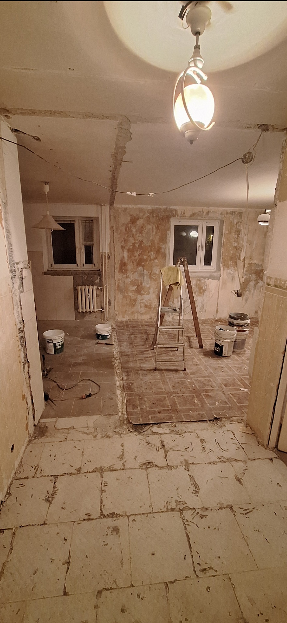 Wnętrze w trakcie remontu: odsłonięte ściany, częściowo zerwana podłoga, drabina i wiadra z farbą. Widoczne okna i lampa sufitowa. Przygotowanie do malowania i wykończenia.