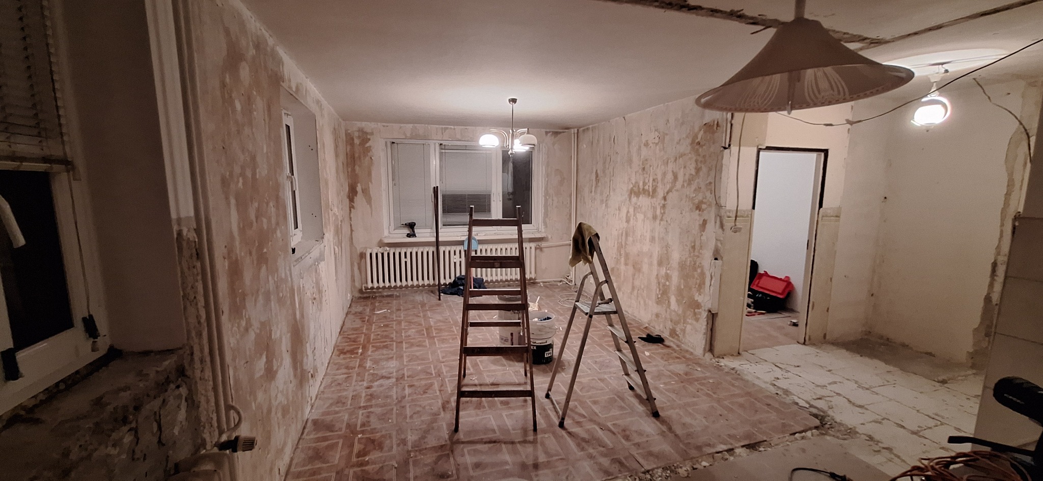 Pokój w trakcie remontu: ściany przygotowane do malowania, widoczne drabiny, narzędzia i resztki starej farby. Podłoga częściowo odsłonięta, widoczne ślady po kleju.