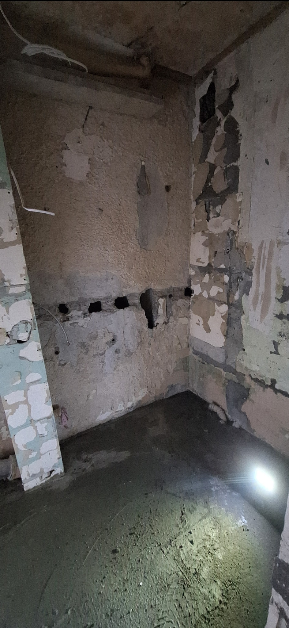 Wnętrze w trakcie remontu: oświetlona podłoga z wylewką, ściany z widocznymi śladami usuniętych płytek i instalacji. Widoczne otwory w ścianie i fragmenty starej farby.